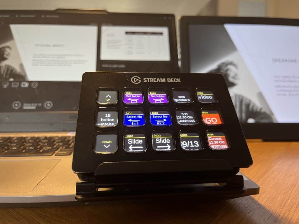 A close-up view of a Stream Deck device on a wooden desk, with colorful buttons labeled for functions like 'Video' and 'Slide.' In the background, there are two screens displaying presentation slides and a document related to a speaking event.