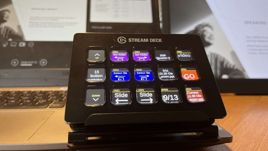 A close-up view of a Stream Deck device on a wooden desk, with colorful buttons labeled for functions like 'Video' and 'Slide.' In the background, there are two screens displaying presentation slides and a document related to a speaking event.