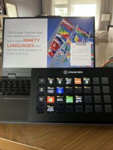 A laptop with a partially visible screen displaying information about the Google Translate app, accompanied by a Stream Deck featuring multiple buttons. Flags of various countries are visible in the background.
