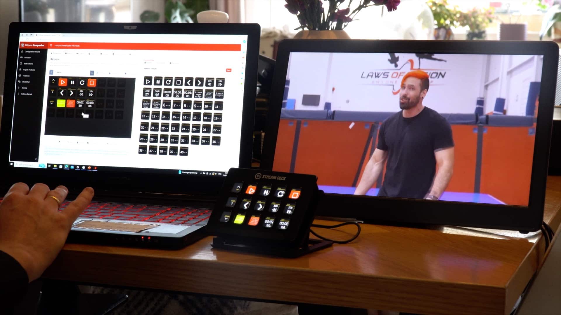 A person's hand on a laptop keyboard, next to a tablet displaying a fitness video featuring a man in a gym. A control panel with buttons is positioned in front of the laptop.