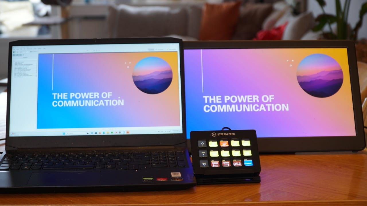 A laptop and an external monitor displaying the text "THE POWER OF COMMUNICATION" over a colorful gradient background. A control pad with colored buttons is shown beside the monitors on a wooden surface.